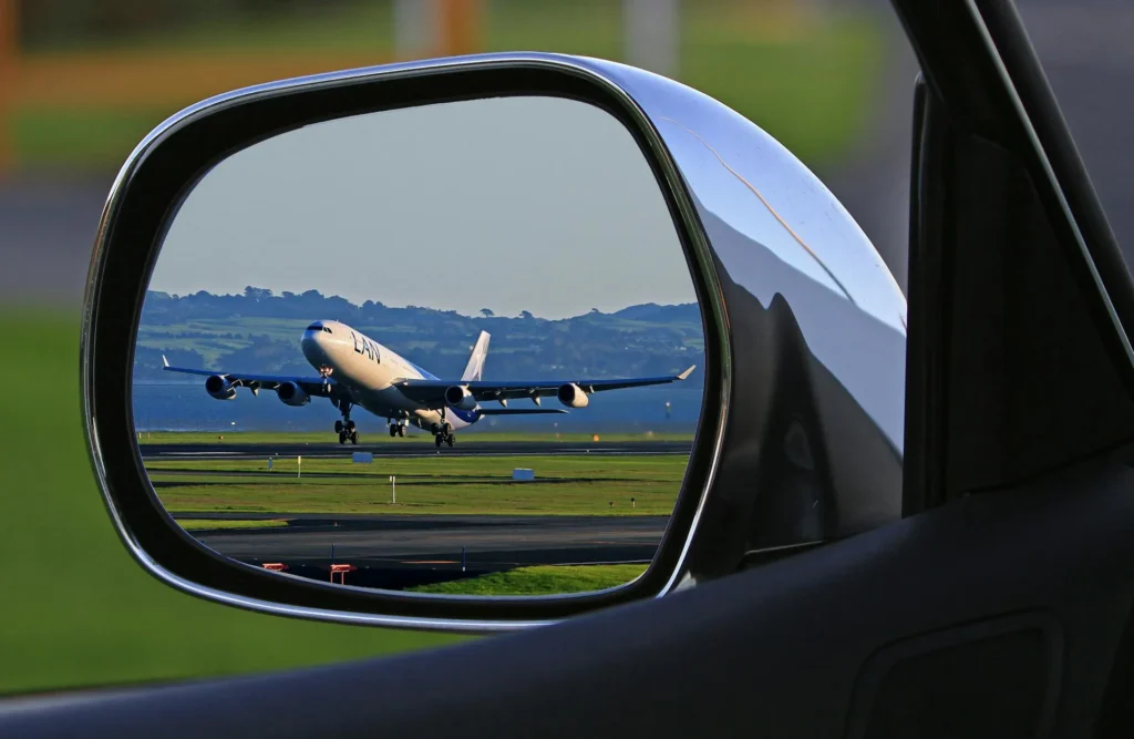 The image shows the rear-view mirror of a car and, in the reflection, a plane about to take off from a runway